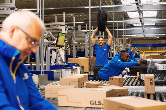 Employees in the depot are sorting packages, one is holding up a car tire and laughing into the camera