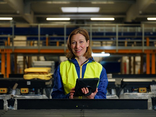 GLS Mitarbeiterin steht mit einem Tablet und Warnweste an der Bandanlage
