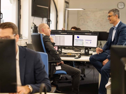 Colleagues in an open plan office talking to each other, one is sitting on the others desk