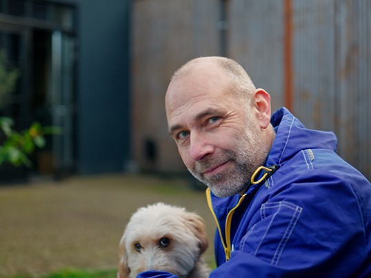 GLS Mitarbeiter mit Arbeitskleidung schaut in die Kamera und neben ihm sitzt ein kleiner Hund